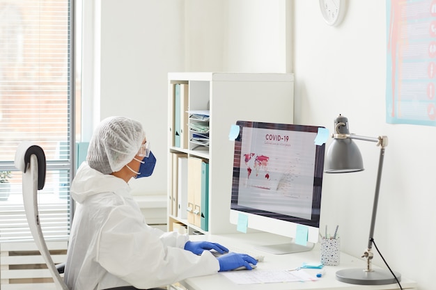 Doctora con ropa protectora sentada en la mesa frente al monitor de la computadora y examinando las estadísticas sobre el coronavirus