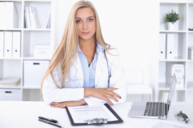 Doctora rubia sentada en la mesa en el hospital y mirando a la cámara.