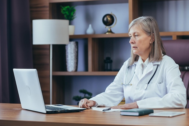 Doctora senior de cabello gris que trabaja en una oficina clásica usando un jefe de computadora portátil de la clínica