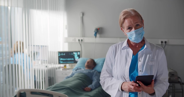 Doctora senior en máscara protectora sosteniendo tableta digital y dando entrevista en cámara