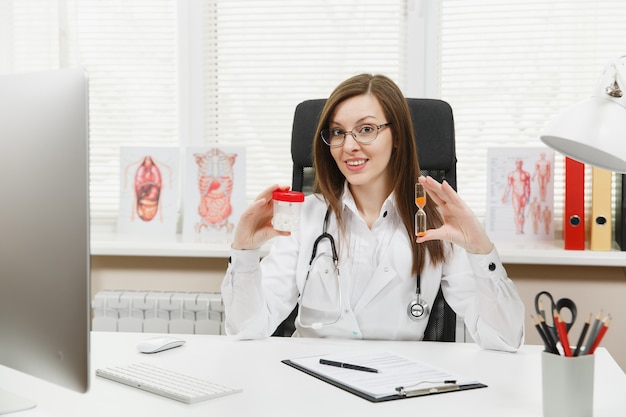 Doctora sentada en el escritorio, sosteniendo una botella con pastillas blancas, reloj de arena, trabajando con documentos médicos en la oficina de luz en el hospital