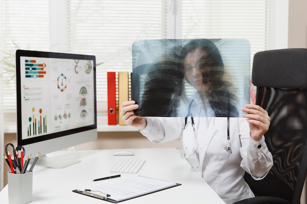 Foto doctora sentada en un escritorio, trabajando en una computadora, sosteniendo una radiografía de los pulmones, fluorografía, roentgen en la oficina de luz en el hospital