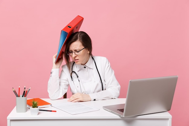 Doctora sentarse en el escritorio de trabajo en la computadora con carpeta de retención de documentos médicos en el hospital aislado sobre fondo de pared rosa pastel. Mujer con estetoscopio de gafas de bata médica. Concepto de medicina sanitaria