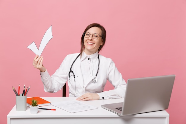 Doctora sentarse en el escritorio de trabajo en la computadora con cheque de retención de documento médico en el hospital aislado sobre fondo de pared rosa pastel. Mujer con estetoscopio de gafas de bata médica. Concepto de medicina sanitaria.