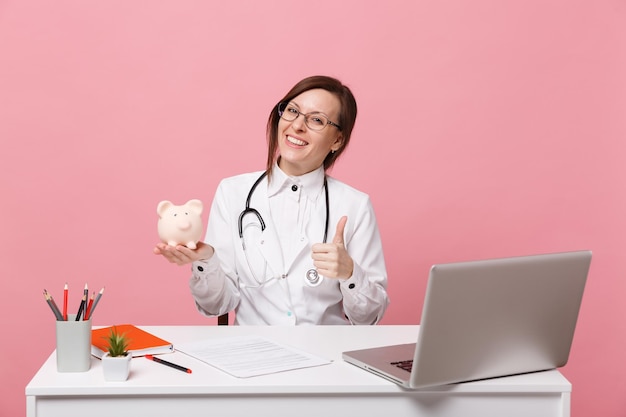 Doctora sentarse en el escritorio de trabajo en la computadora con documento médico mantenga dinero de cerdo en el hospital aislado sobre fondo rosa pastel. Mujer con estetoscopio de gafas de bata médica. Concepto de medicina sanitaria.