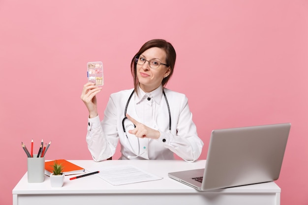 Doctora sentarse en el escritorio de trabajo en la computadora con documento médico mantenga píldoras en el hospital aislado sobre fondo de pared rosa pastel. Mujer con estetoscopio de gafas de bata médica. Concepto de medicina sanitaria.