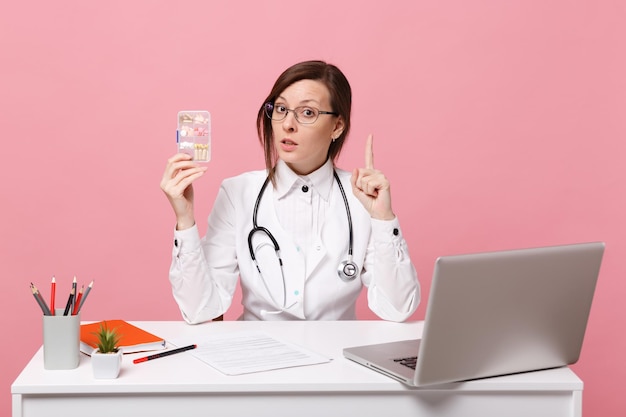 Doctora sentarse en el escritorio de trabajo en la computadora con documento médico mantenga píldoras en el hospital aislado sobre fondo de pared rosa pastel. Mujer con estetoscopio de gafas de bata médica. Concepto de medicina sanitaria.
