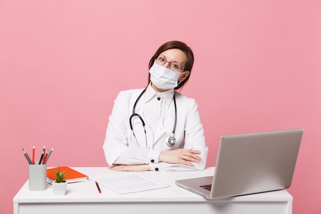 Doctora sentarse en el escritorio de trabajo en computadora con documento médico en mascarilla en hospital aislado sobre fondo de pared rosa pastel. Mujer en bata médica gafas estetoscopio concepto de medicina sanitaria