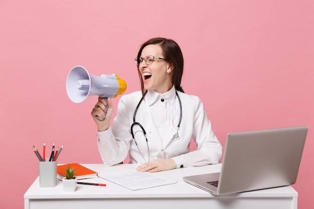 Doctora sentarse en el escritorio de trabajo en la computadora con documento médico sostenga el megáfono en el hospital aislado sobre fondo rosa pastel. Mujer con estetoscopio de gafas de bata médica. Concepto de medicina sanitaria.