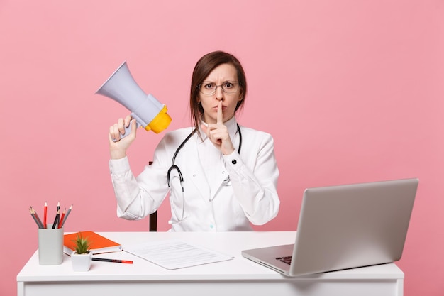 Doctora sentarse en el escritorio de trabajo en la computadora con documento médico sostenga el megáfono en el hospital aislado sobre fondo rosa pastel. Mujer con estetoscopio de gafas de bata médica. Concepto de medicina sanitaria.