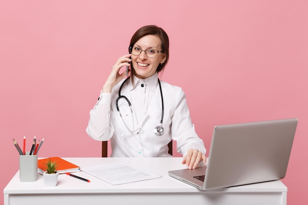 Doctora sentarse en el escritorio de trabajo en la computadora con documento médico sostenga el teléfono celular en el hospital aislado sobre fondo rosa pastel. Mujer con estetoscopio de gafas de bata médica. Concepto de medicina sanitaria.