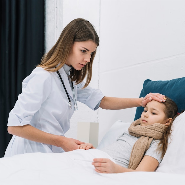 Doctora seria que controla la temperatura de una niña acostada en la cama con fiebre