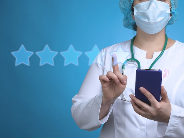 Una doctora se para sobre una superficie azul y sostiene un teléfono inteligente, con la otra mano hace clic en las estrellas de calificación virtuales