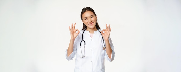 Una doctora sonriente, una trabajadora de la salud asiática con uniforme de clínica médica, muestra signos de aprobación, algo que da permiso al paciente, se encuentra sobre fondo blanco.