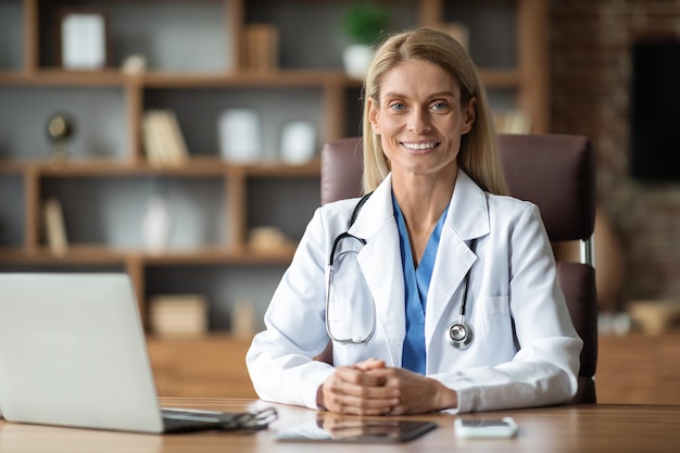 Doctora sonriente en uniforme sentada en el escritorio con una computadora portátil en el lugar de trabajo