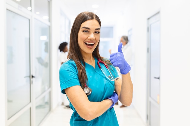 Doctora sonriente vistiendo matorrales en el concurrido pasillo del hospital mostrando los pulgares para arriba