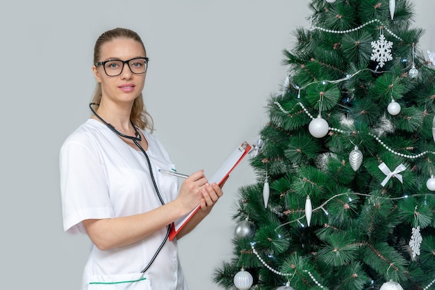 Doctora sostiene un portapapeles en el fondo de un árbol de Navidad. Concierte una cita en el hospital