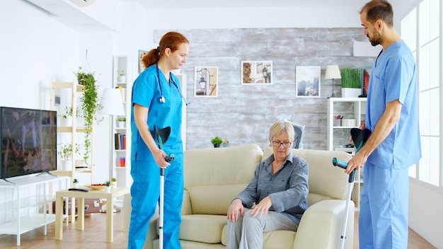 Doctora y su asistente ayudando a una anciana con muletas a levantarse del sofá y dar un paseo