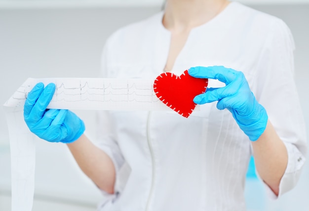 La doctora tiene un corazón rojo y una copia impresa en papel de un cardiograma. Prevención de enfermedades cardiovasculares.