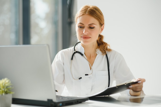 Doctora trabajando en el escritorio de oficina y sonriendo a la cámara, interior de la oficina en el fondo