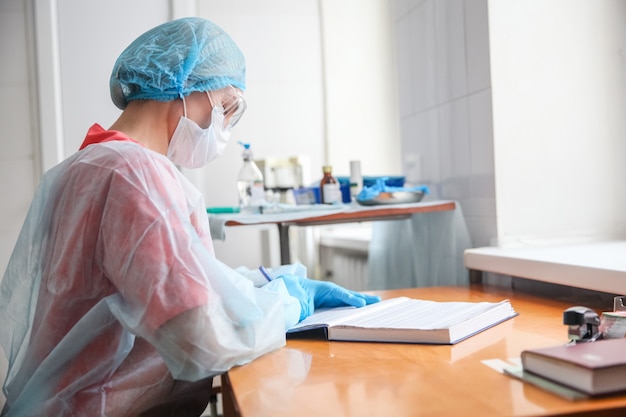 Una doctora con un traje de protección blanco, gorra azul, guantes azules y máscara se sienta en una mesa y llena una revista