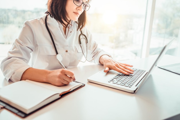 Doctora en uniforme blanco con estetoscopio en el cuello en el papeleo con cuaderno y computadora portátil