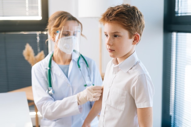 Doctora con uniforme blanco y máscara de seguridad aplicando yeso en el hombro del niño después de la inyección de vacunación en el hospital