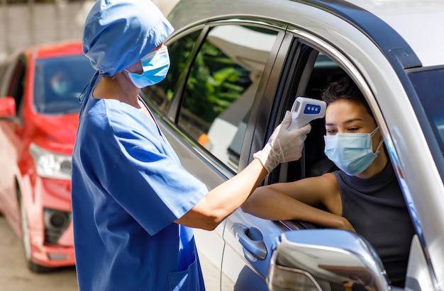 Doctora en uniforme de hospital azul y soporte de mascarilla cerca de la unidad a través de la cola del automóvil.