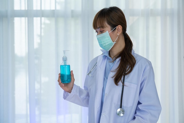 Doctora usa uniforme blanco y mascarilla sosteniendo una botella de alcohol en gel