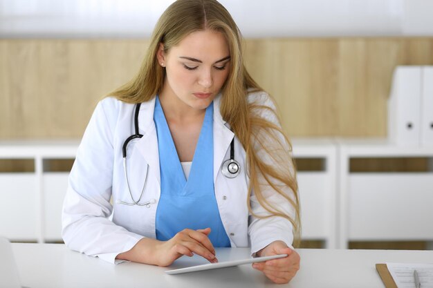 Doctora usando tablet pc en hospital o clínica Terapeuta rubia sonriendo alegremente Datos en medicina y concepto de salud