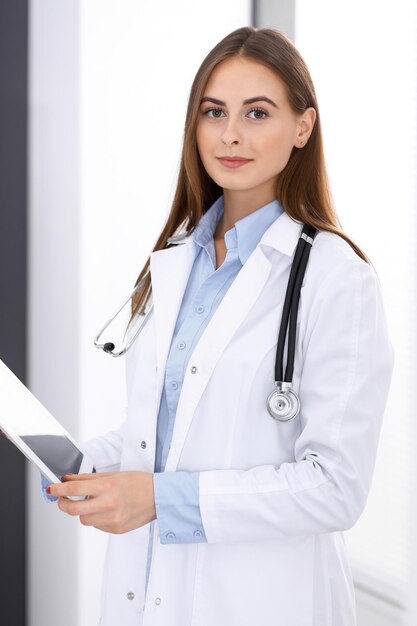 Doctora usando una tableta mientras está de pie cerca de la ventana en el hospital. Médico feliz en el trabajo. Concepto de medicina y atención de la salud.