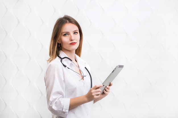 Doctora usando tableta mujer inteligente y encantadora con el aparato y el estetoscopio en el fondo blanco