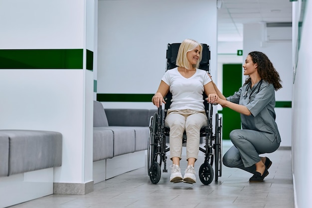 Una doctora vestida con un traje médico sostiene la mano de una mujer en silla de ruedas en el pasillo del hospital