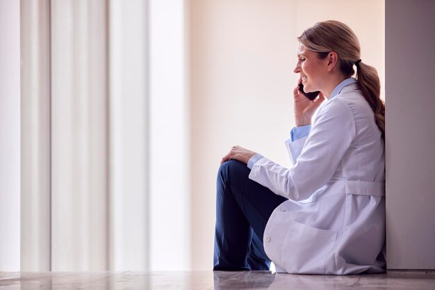 Doctora vistiendo bata blanca sentada en el suelo en el pasillo del hospital haciendo llamadas en el teléfono móvil