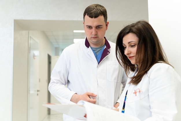 Doctores masculinos y femeninos hablando y mirando los papeles en la clínica. Hombre y mujer, médicos que tienen discusión en el hospital. Médico y enfermera discutiendo la enfermedad. Conversación con el asistente y los documentos