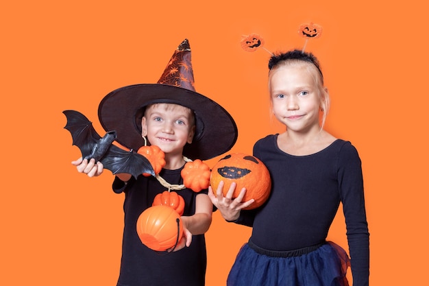 Doçura ou travessura infantil em uma fantasia de Halloween. Menino com chapéu de mago segurando uma cesta laranja em forma de abóbora e um morcego preto e uma menina segurando uma abóbora pintada em um fundo laranja.