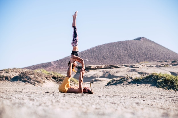 Dois adultos fazendo acroyoga juntos na praia na areia - um homem segurando seu companheiro de equipe com as pernas