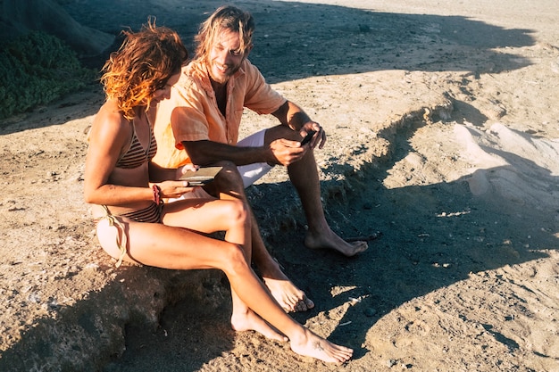 Dois adultos na praia conversando e lambendo o telefone da mulher sentada nas pedras - mulher de biquíni olhando para o telefone e um homem olhando para o mesmo telefone