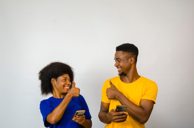 Dois africanos fofos lado a lado isolados sobre fundo branco sorriem enquanto operam seu celular e fazem polegares para cima