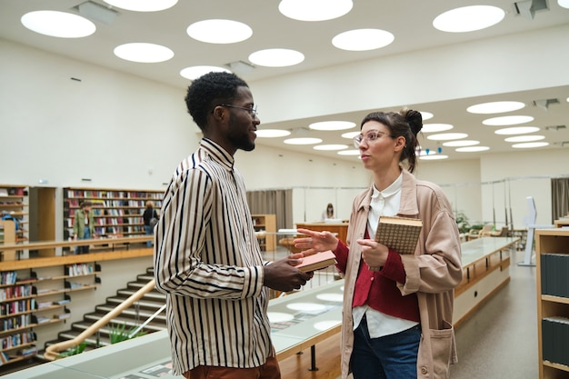 Dois alunos segurando livros e discutindo algo enquanto estudavam na biblioteca