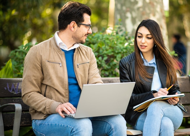 Dois alunos sentados em um banco ao ar livre usando livros e um laptop