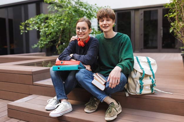 Dois alunos sorridentes sentados com pastas e livros nas mãos e felizes no pátio da universidade