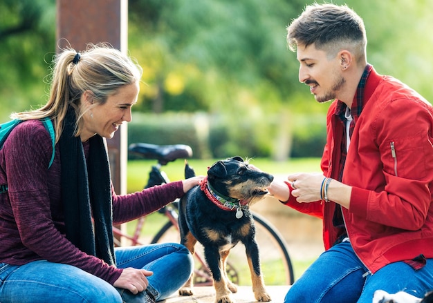 Dois amigos acariciando um cachorrinho sentado em uma parede de tijolos de um parque público