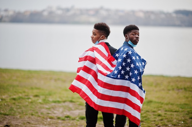 Dois amigos africanos de adolescentes com a bandeira americana no parque usando máscaras médicas protegem da quarentena do vírus de coronavírus de infecções e doenças.