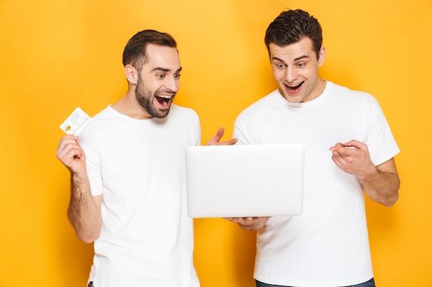 Dois amigos alegres e animados vestindo camisetas em branco, isolados na parede amarela, usando um laptop, comemorando o sucesso, mostrando um cartão de crédito de plástico