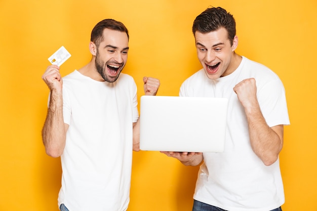 Dois amigos alegres e animados vestindo camisetas em branco, isolados na parede amarela, usando um laptop, comemorando o sucesso, mostrando um cartão de crédito de plástico