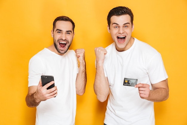 Dois amigos alegres e entusiasmados usando camisetas em branco, isolados sobre uma parede amarela, olhando para o celular, comemorando o sucesso