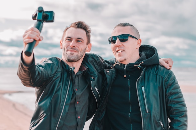 Dois amigos bonitos e sorridentes fazendo selfie usando a câmera de ação com estabilizador de cardan na praia.