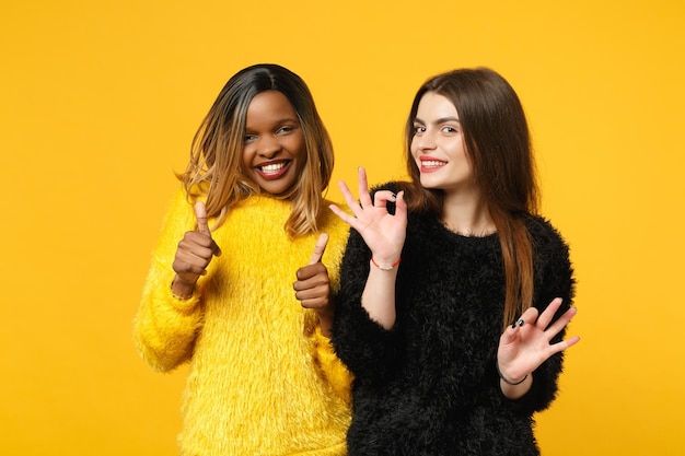 Dois amigos de mulheres jovens europeus e afro-americanos em roupas amarelas pretas em pé posando isolado no fundo da parede laranja brilhante, retrato de estúdio. conceito de estilo de vida de pessoas. mock up espaço de cópia.