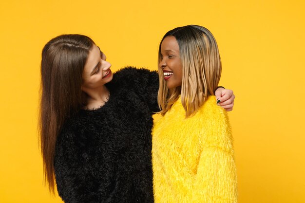 Dois amigos de mulheres jovens europeus e afro-americanos em roupas amarelas pretas em pé posando isolado no fundo da parede laranja brilhante, retrato de estúdio. Conceito de estilo de vida de pessoas. Mock up espaço de cópia.